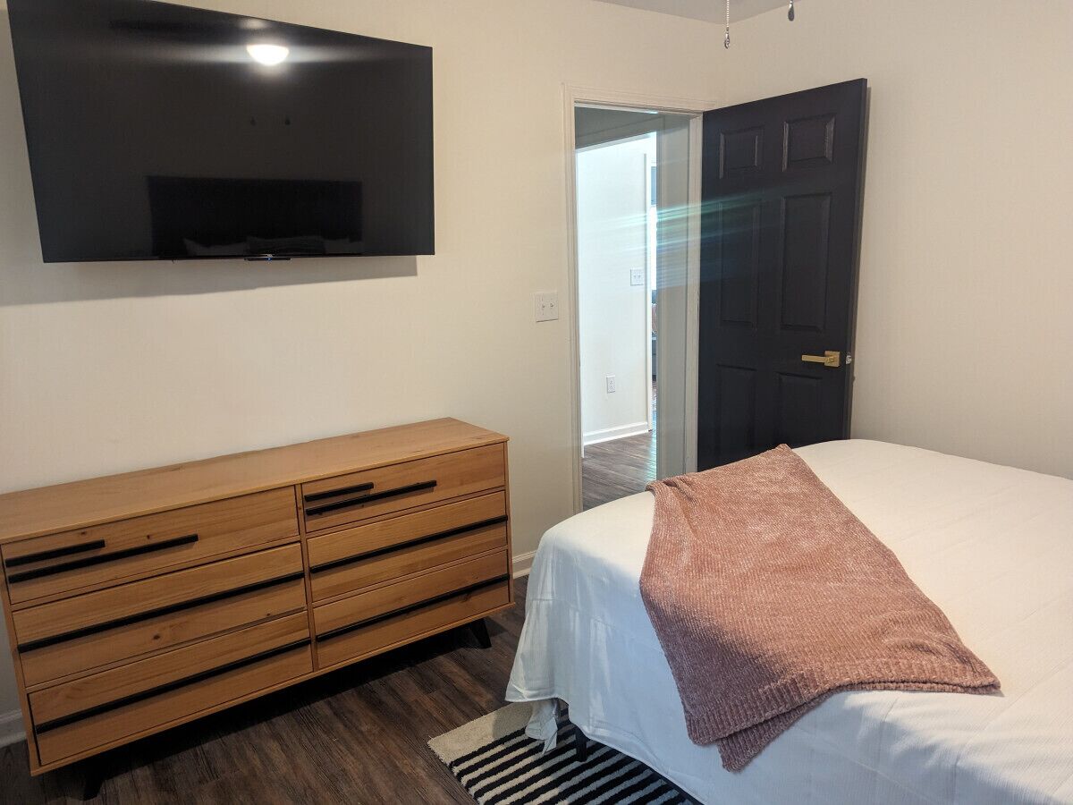 Quarto com cama, cômoda de madeira, televisão na parede e porta que dá para outro espaço.