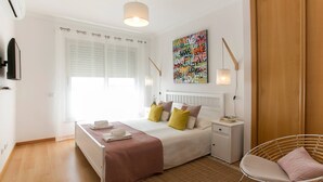 Een moderne slaapkamer met een bed, nachtkastjes, een stoel, een raam met jaloezieën, een kunstwerk aan de muur en een hanglamp.
