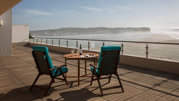Een balkon met twee stoelen en een tafel, met uitzicht op een strand met golven en veraf gelegen land.