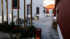 A courtyard with a table and chairs, a potted plant, and a building with red shutters and a tiled roof. - Casa Flamingo - Casa Principal, Foz do Arelho (Caldas da Rainha)