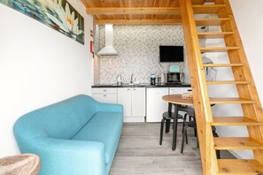 Un espace de vie compact comprenant un coin cuisine, un canapé bleu, une échelle en bois et un sol carrelé.