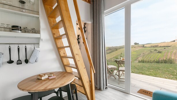 Ein modernes Interieur mit einem Holztisch, Stühlen, einer Leiter und Blick auf einen Weinberg durch große Fenster.