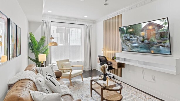 Una sala moderna con un sofá de cuero, un televisor de pantalla plana montado en la pared, un escritorio con una silla y un amplio ventanal con cortinas.