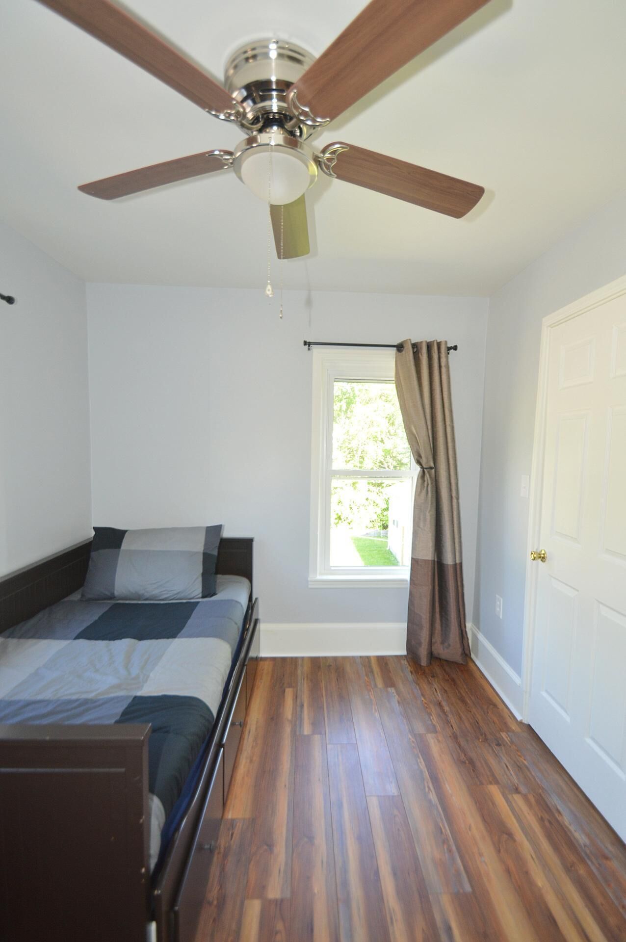 Une chambre à coucher avec un lit, un ventilateur de plafond, une fenêtre avec des rideaux et une porte.