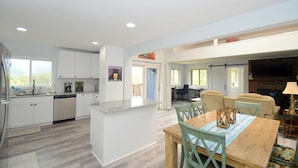A spacious kitchen with white cabinets, a granite countertop, and a central island. There is a dining area with a wooden table and green chairs. A fireplace is visible in the background. - Black Bear Cabin - The Mountain Retreat! (Berkeley Springs)