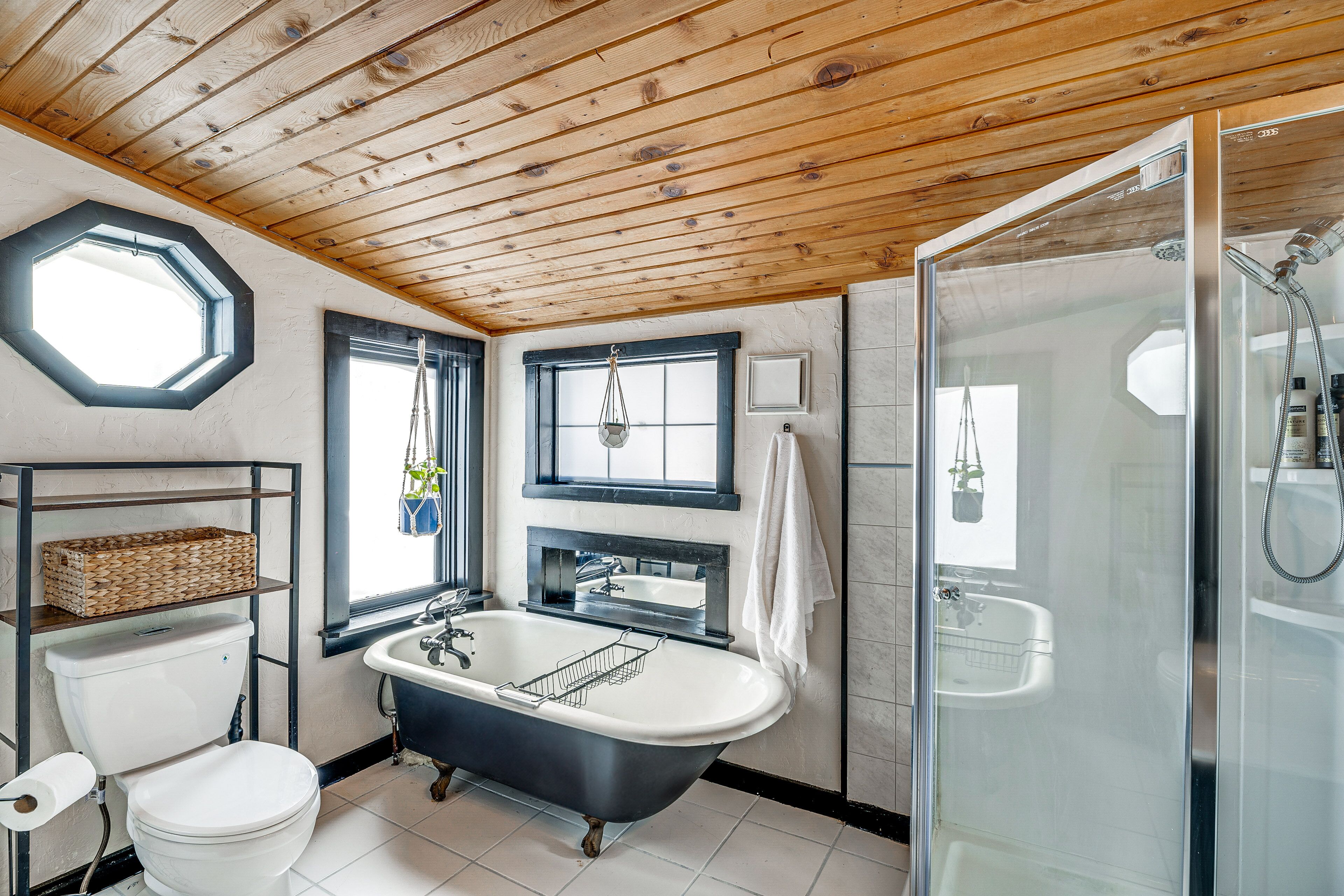 Une salle de bain avec une baignoire sur pieds, une douche à parois de verre, des toilettes et une fenêtre ornée d’une étagère murale.