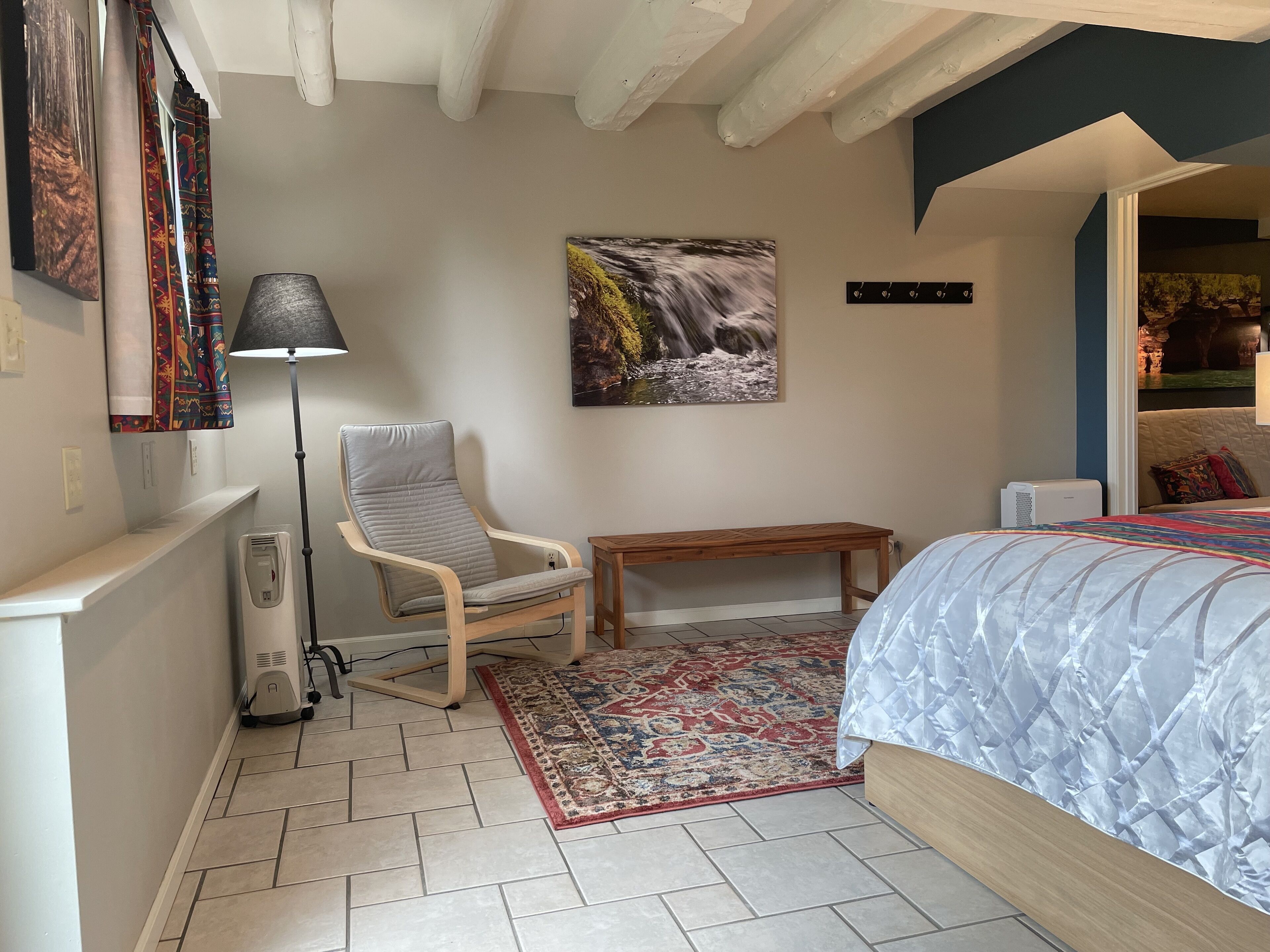 Un dormitorio con una cama, una silla, un banco, una alfombra, un cuadro y un calentador.