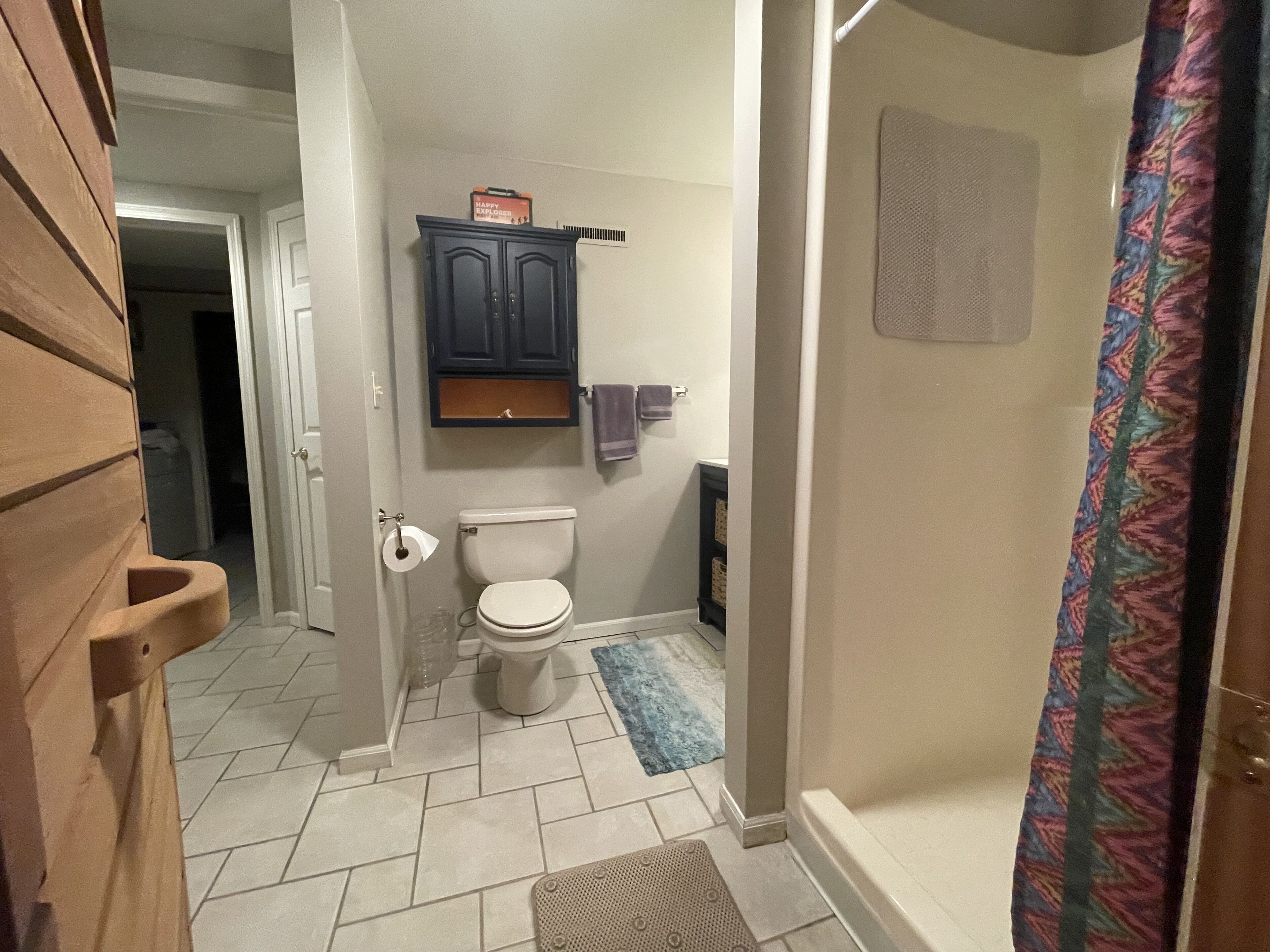 A bathroom with a toilet, a shower with a patterned curtain, and a small cabinet above the toilet.