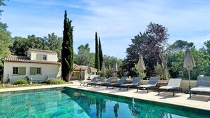 A pool area with lounge chairs and umbrellas, surrounded by trees and a building with a tiled roof. - Lovely home in Nans-Les-Pins (Nans-Les-Pins)