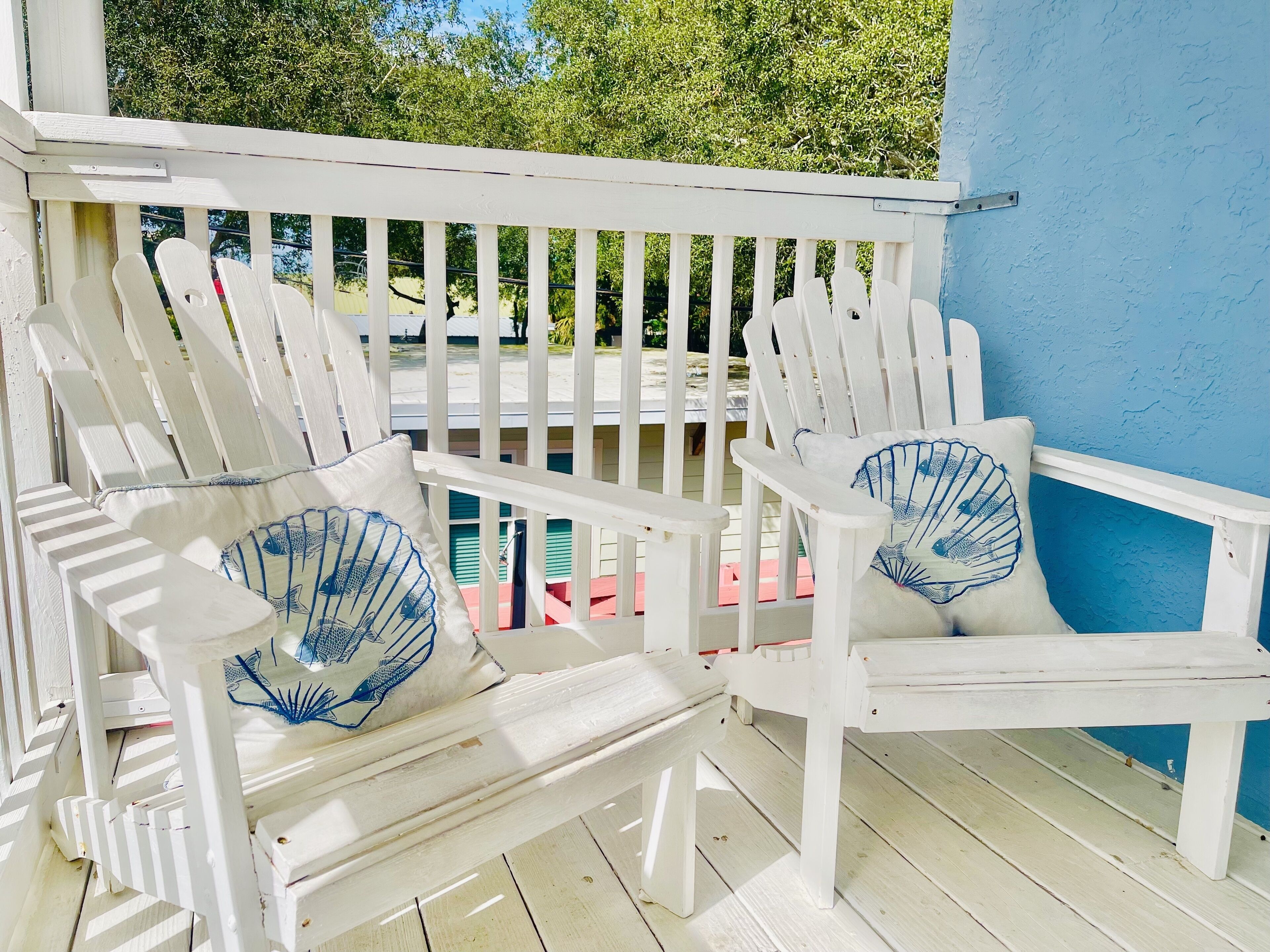 A white wooden porch with two Adirondack chairs, each with a blue fish design pillow.
