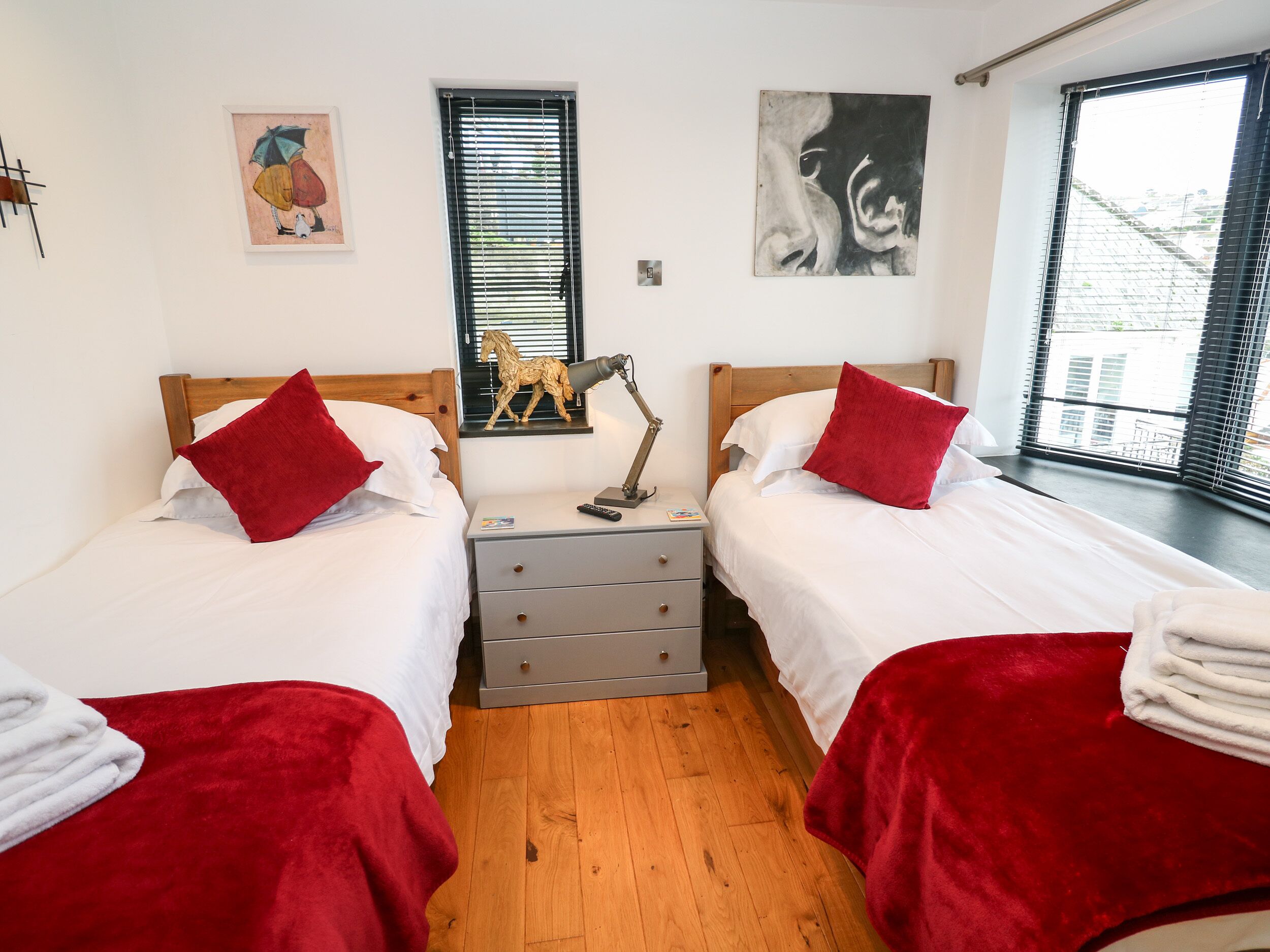 A double room with two beds, each with a red pillow and white bedding. There is a wooden headboard, a bedside table with a lamp, and a small shelf with a horse figurine. The room has a window with blinds, a painting on the wall, and a view of buildings outside.