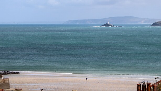 A coastal beach scene with a lighthouse on a small island, calm sea, and a sandy shore. - Rooftops (St Ives)