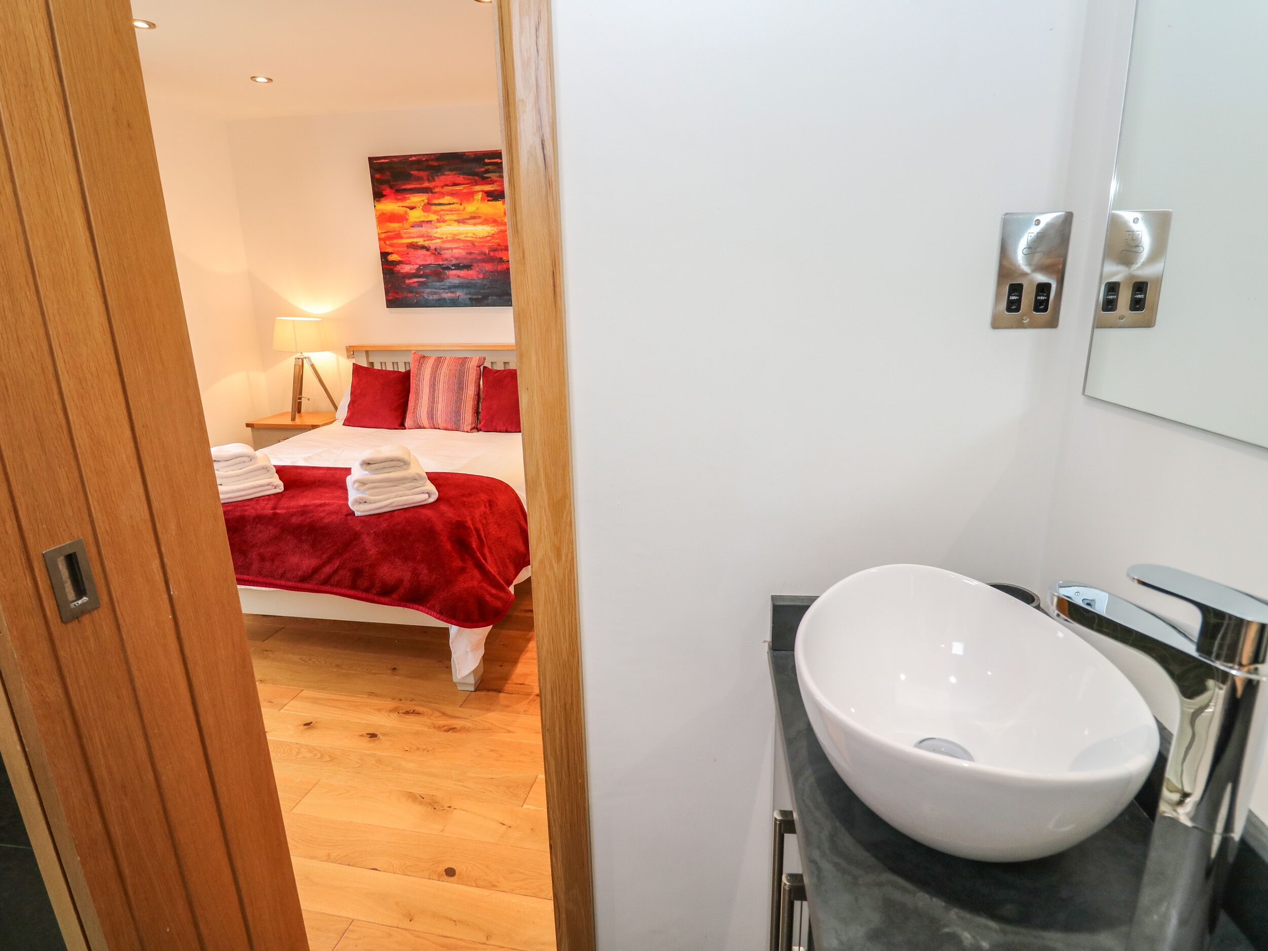 A hotel room with a bed, a red blanket, a white sink, and a painting on the wall.