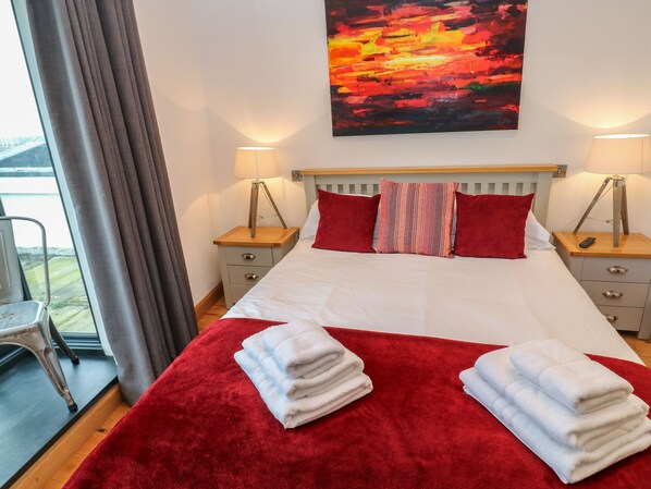 A hotel room with a large bed, two bedside tables with lamps, a red throw blanket, and a painting of a sunset on the wall.