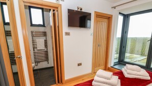 A modern bedroom with a wooden floor, a large bed with a red blanket, a TV on the wall, and a view of the outside through a large window. - Rooftops (St Ives)
