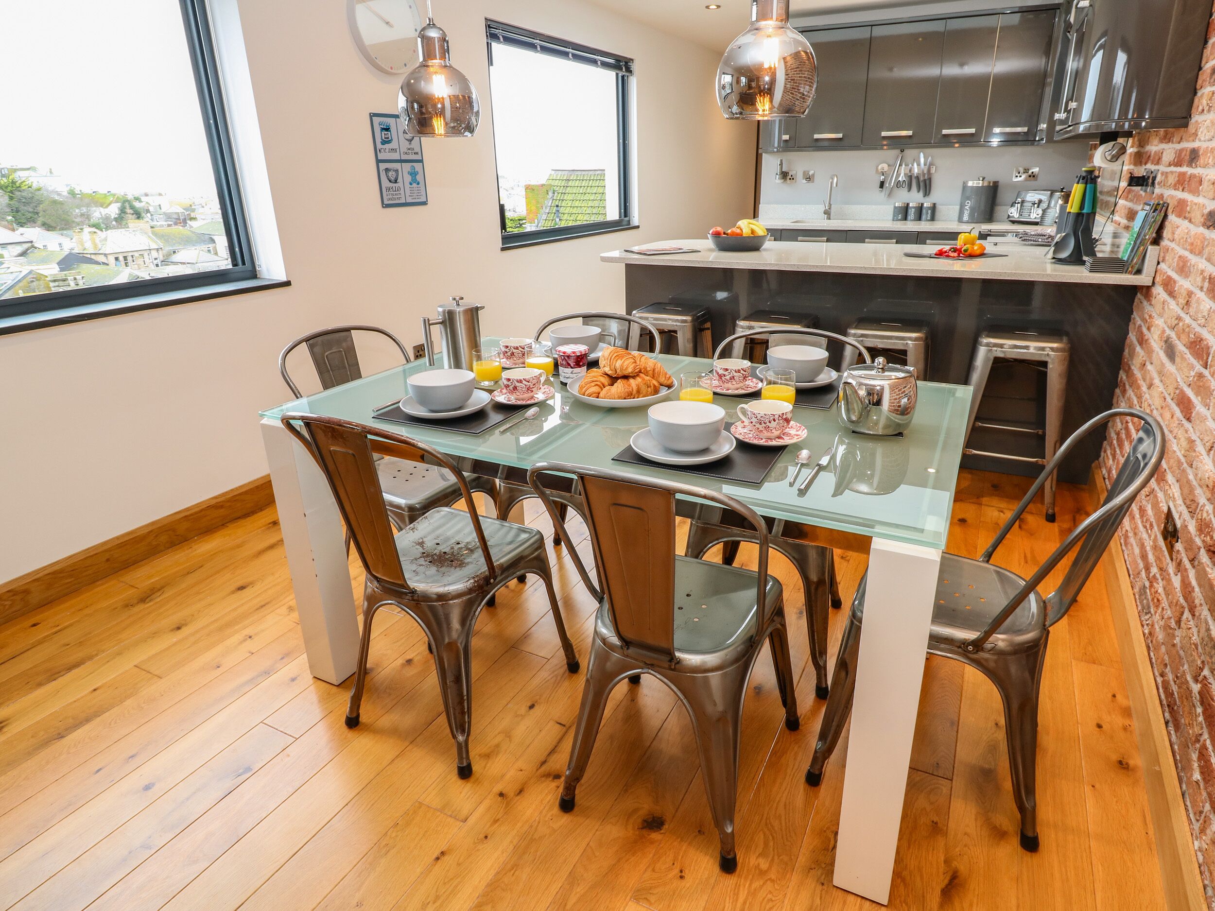 Une cuisine moderne avec un coin repas, comprenant une table en verre, des chaises en métal et un mur en briques.