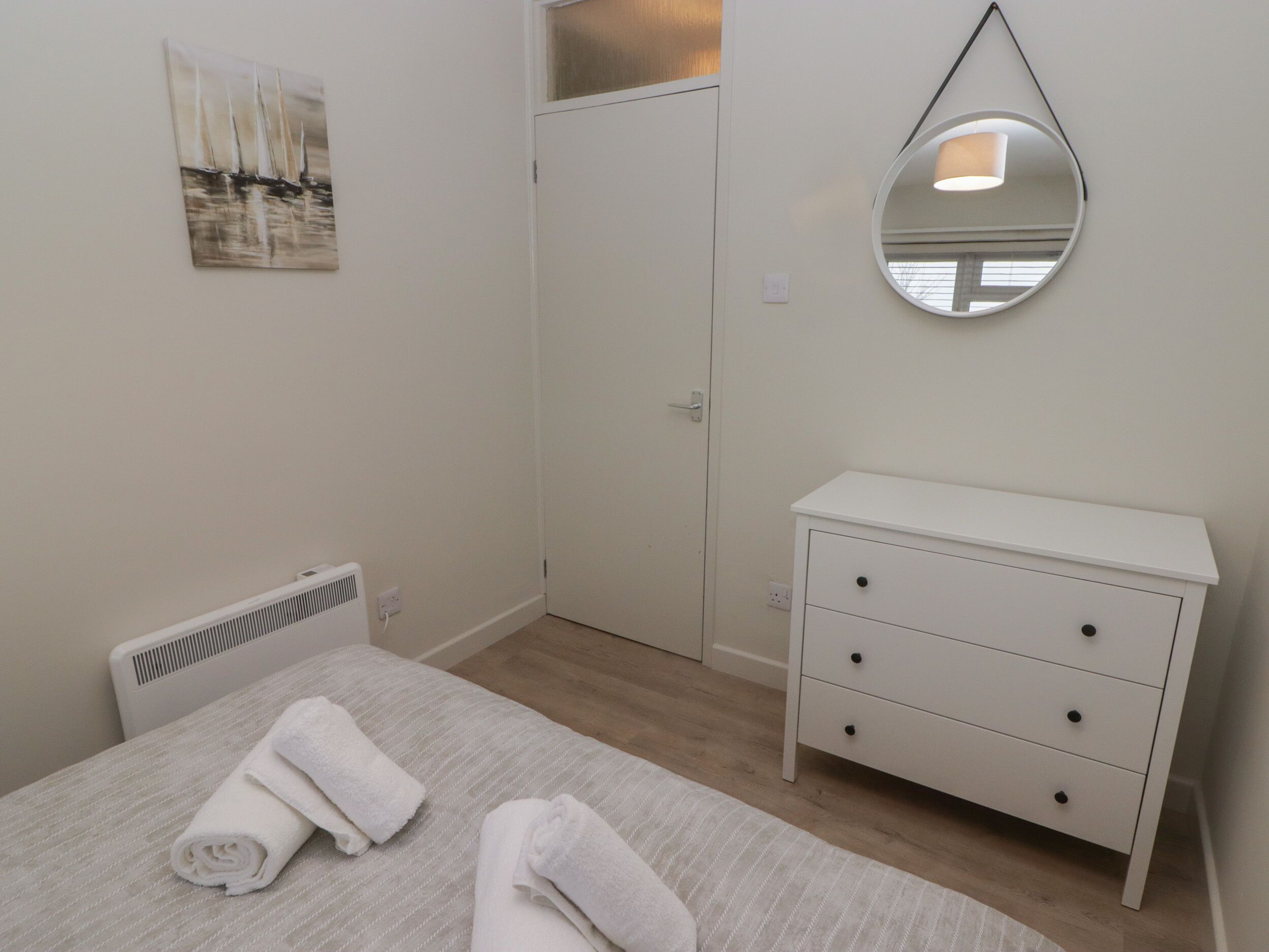 A bedroom with a bed, a bedside table with drawers, a radiator, a door, and a wall-mounted mirror.