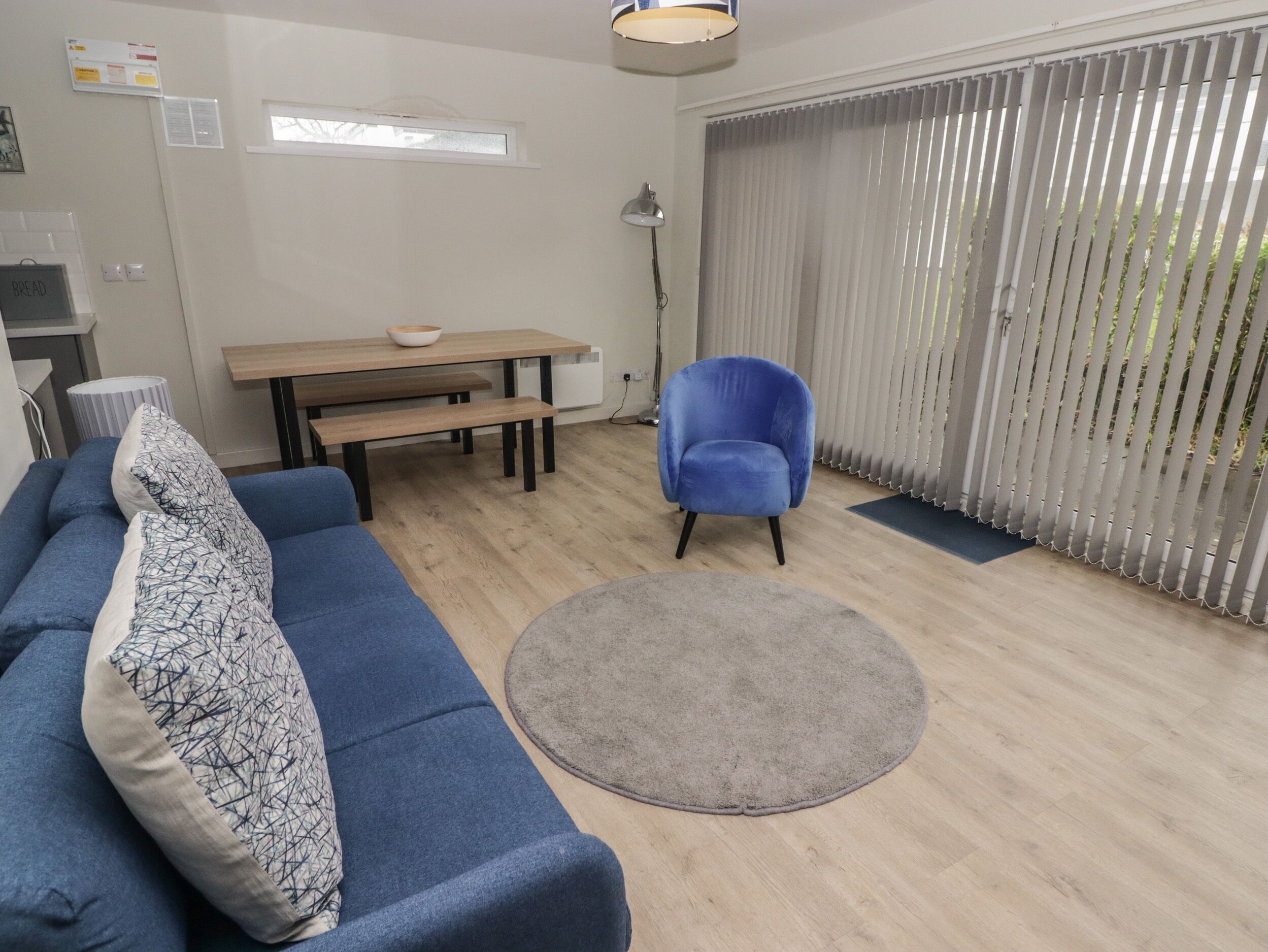A living room with a blue sofa, a round rug, a wooden table with benches, a blue armchair, and a window with blinds.