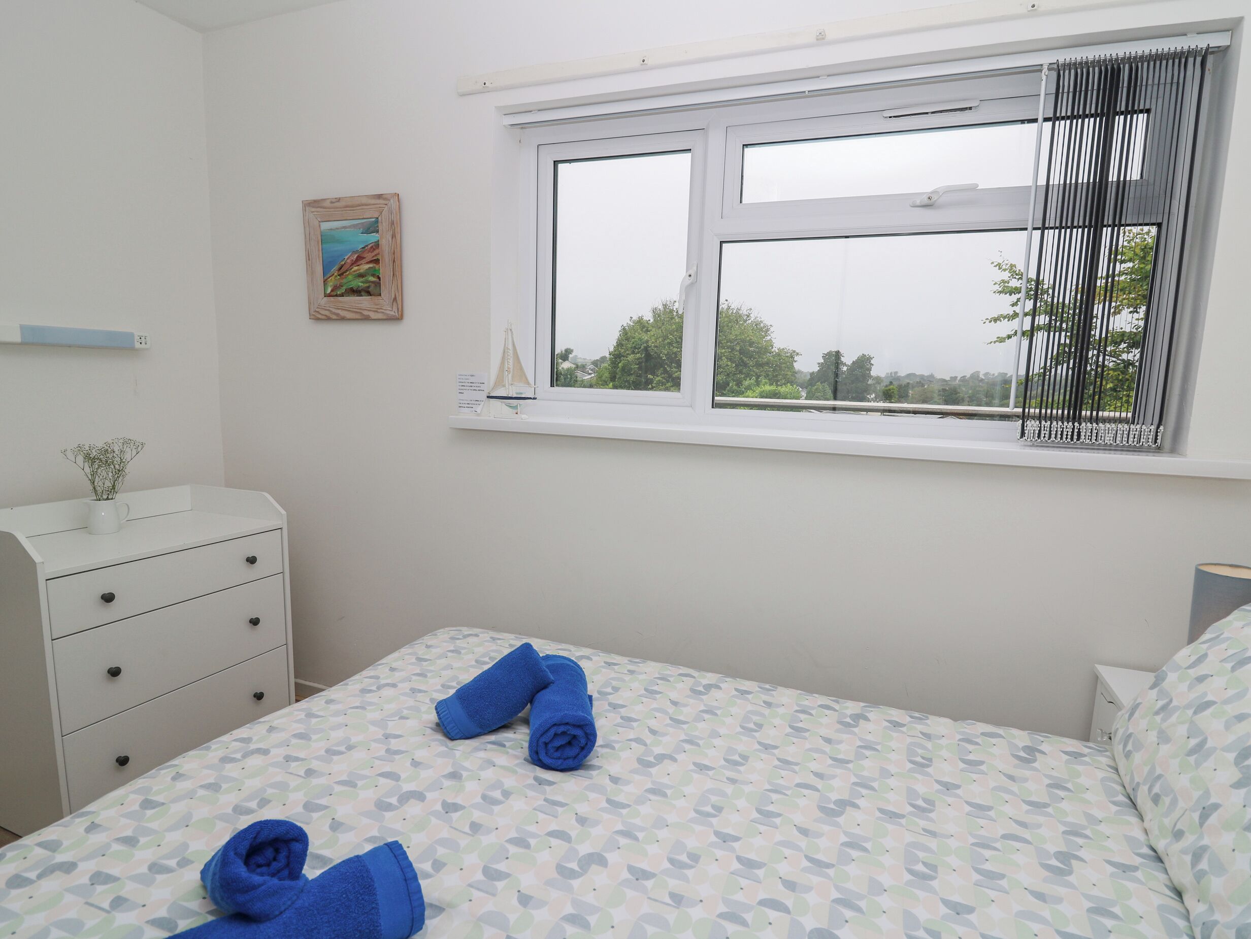 Ein Schlafzimmer mit Bett, Nachttisch, Kommode, einem Fenster mit Aussicht und einem kleinen Bild an der Wand.
