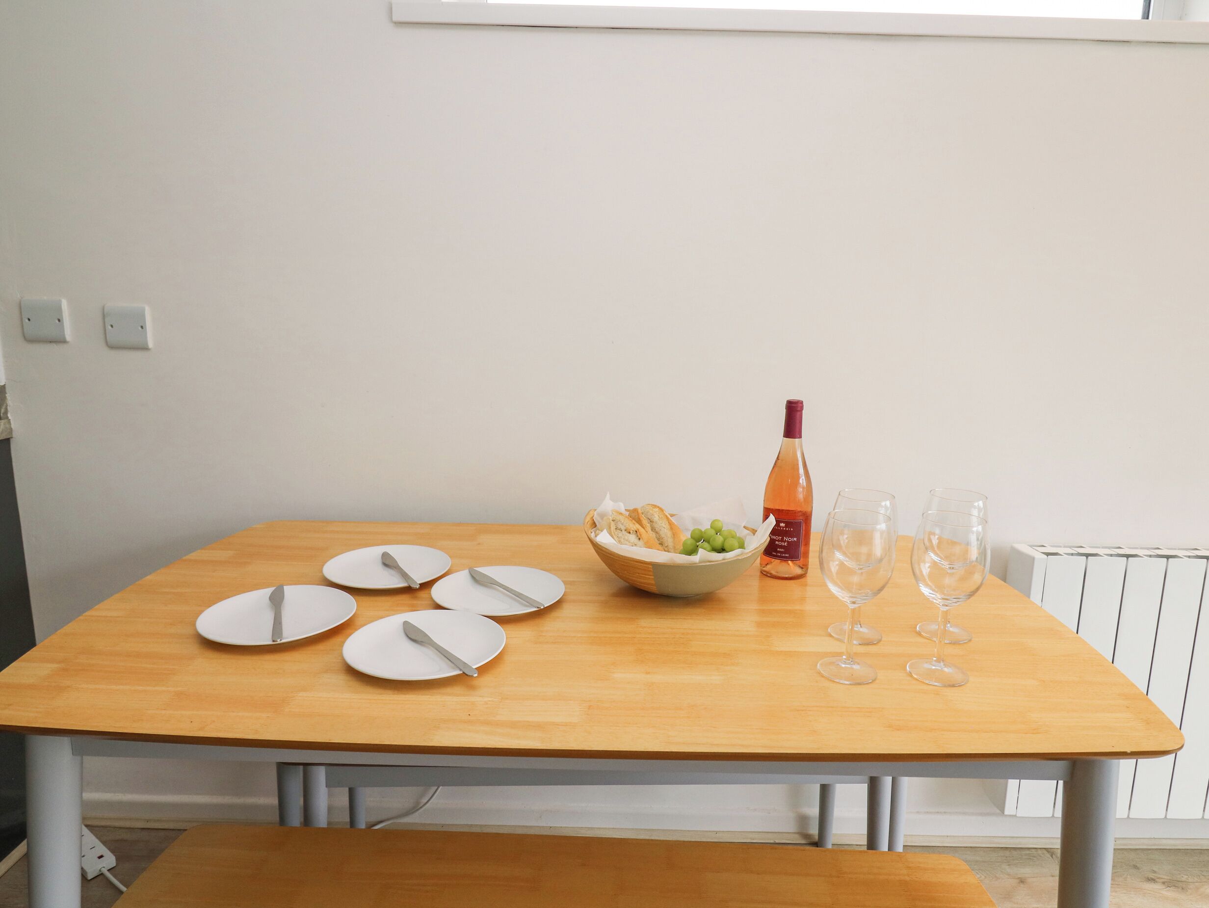 A wooden dining table with white plates, a bowl of grapes, a bottle of wine, and two wine glasses.
