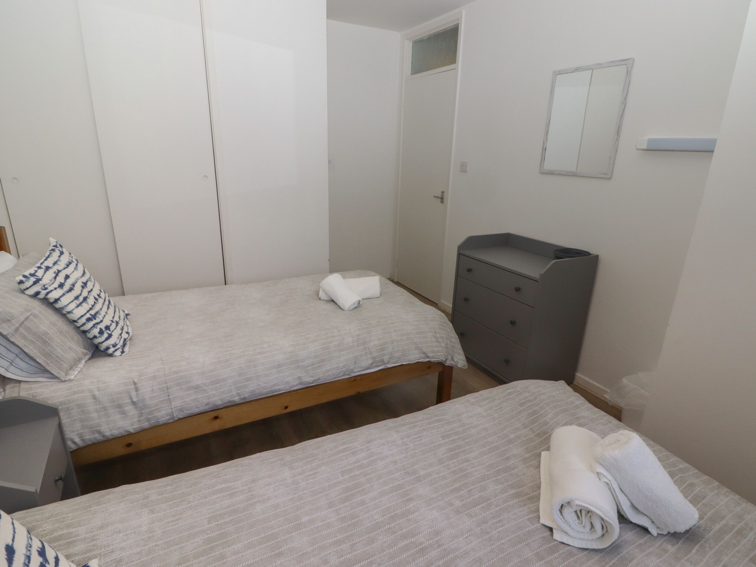 Ett litet, rent rum med två enkelsängar, en grå byrå och en vit garderob.