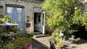 Une charmante maison à la façade en briques, avec une porte en bois et un petit jardin agrémenté de plantes en pot et d’une mangeoire à oiseaux.