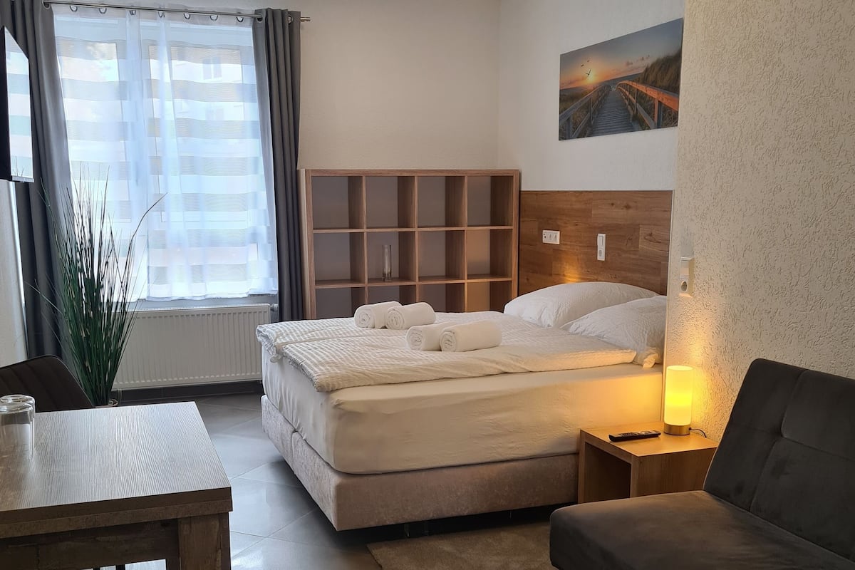 A modern hotel room with a bed, a sofa, a dining table, and a large window with curtains.