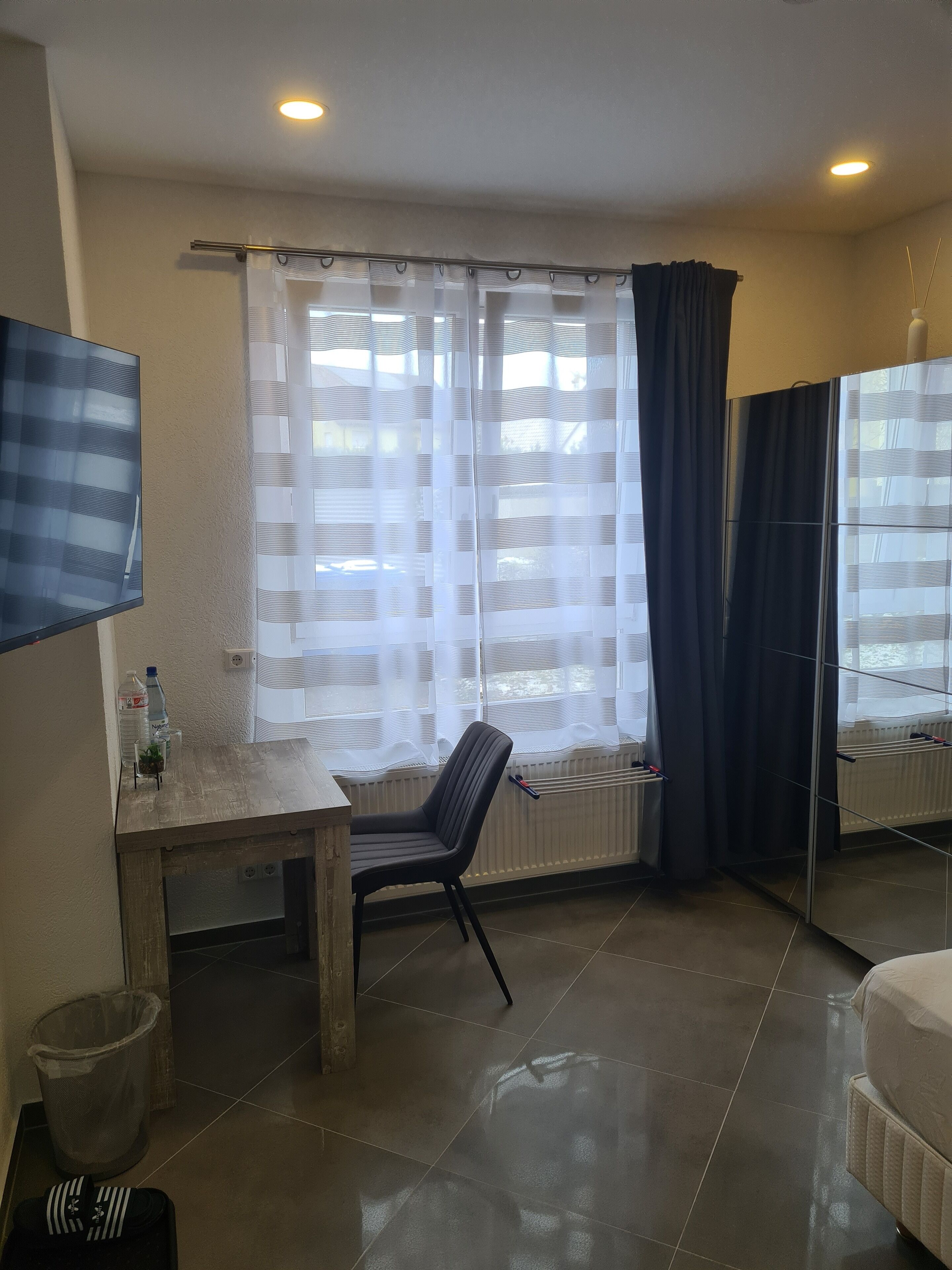 Una habitació d'hotel amb un llit, un escriptori amb una cadira, un televisor fixat a la paret, un radiador i una finestra amb cortines.