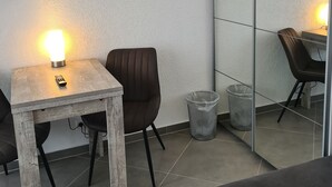 A small, modern room with a wooden table, a single brown chair, a trash bin, and a large mirror on the wall. - WellandHome - 6 rooms (Aalen)