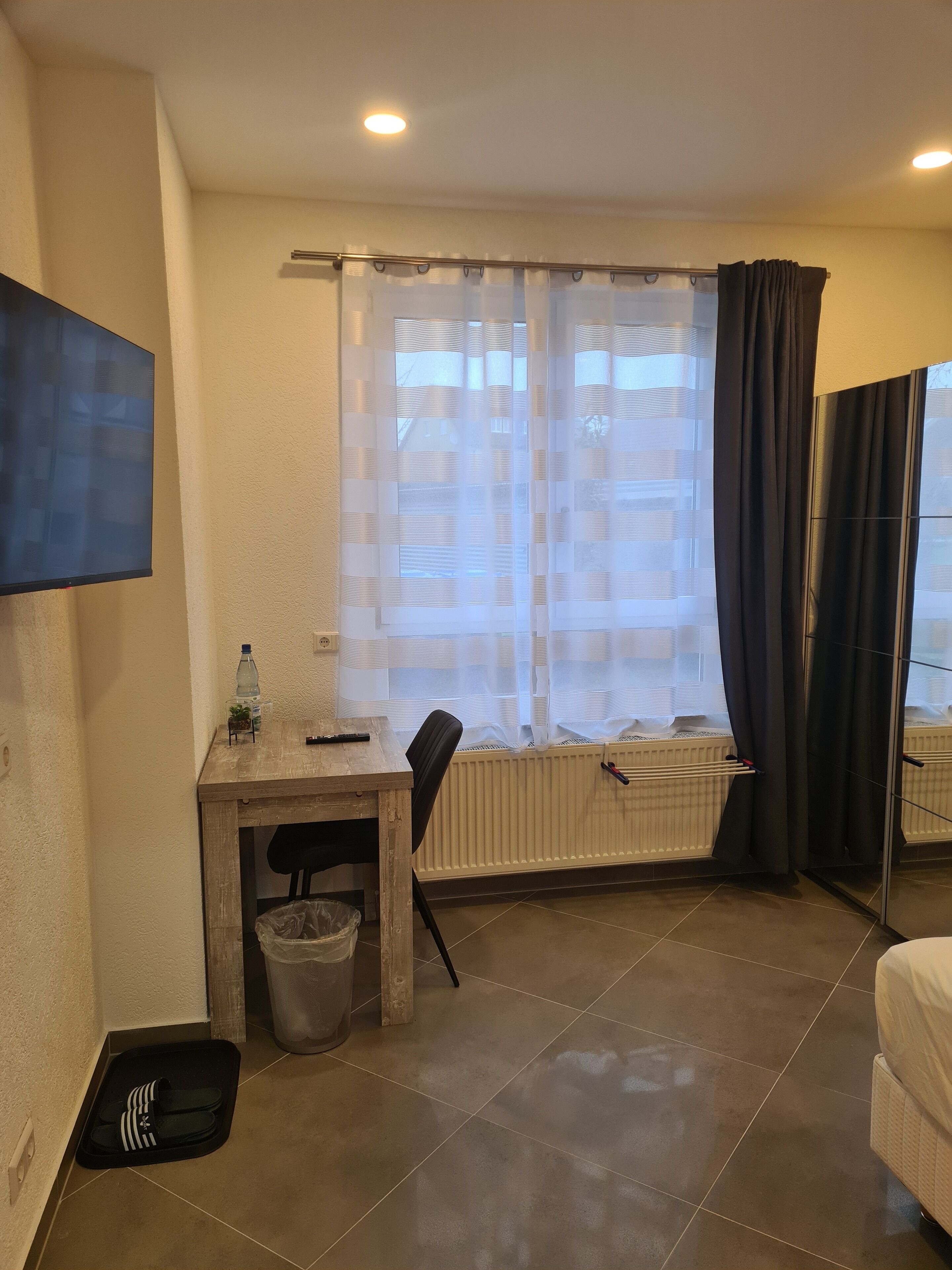 Ein Hotelzimmer mit einem Holztisch, einem Sessel, einem an der Wand montierten Fernseher, einer Heizung, einem Fenster mit leichtem Vorhang und einem Bett.