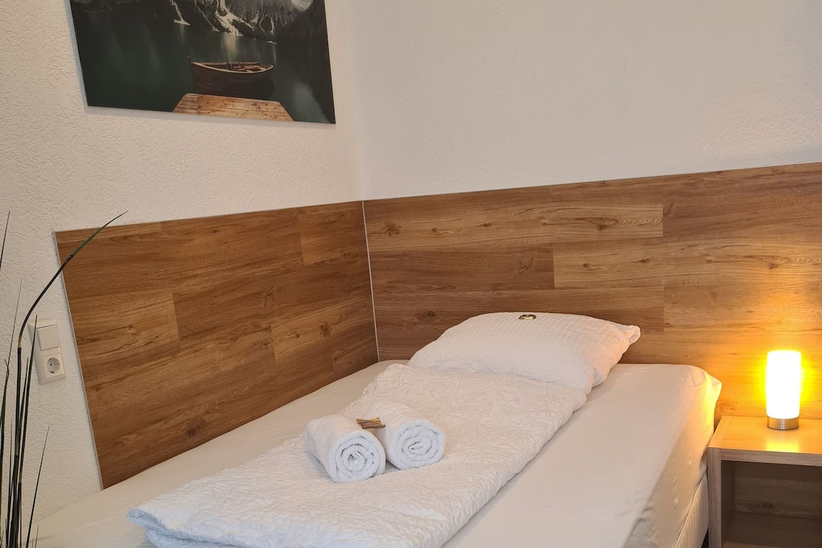 A neatly made bed with white linens and two rolled towels, a wooden headboard, a bedside table with a lamp, and a painting of a mountainous landscape on the wall.