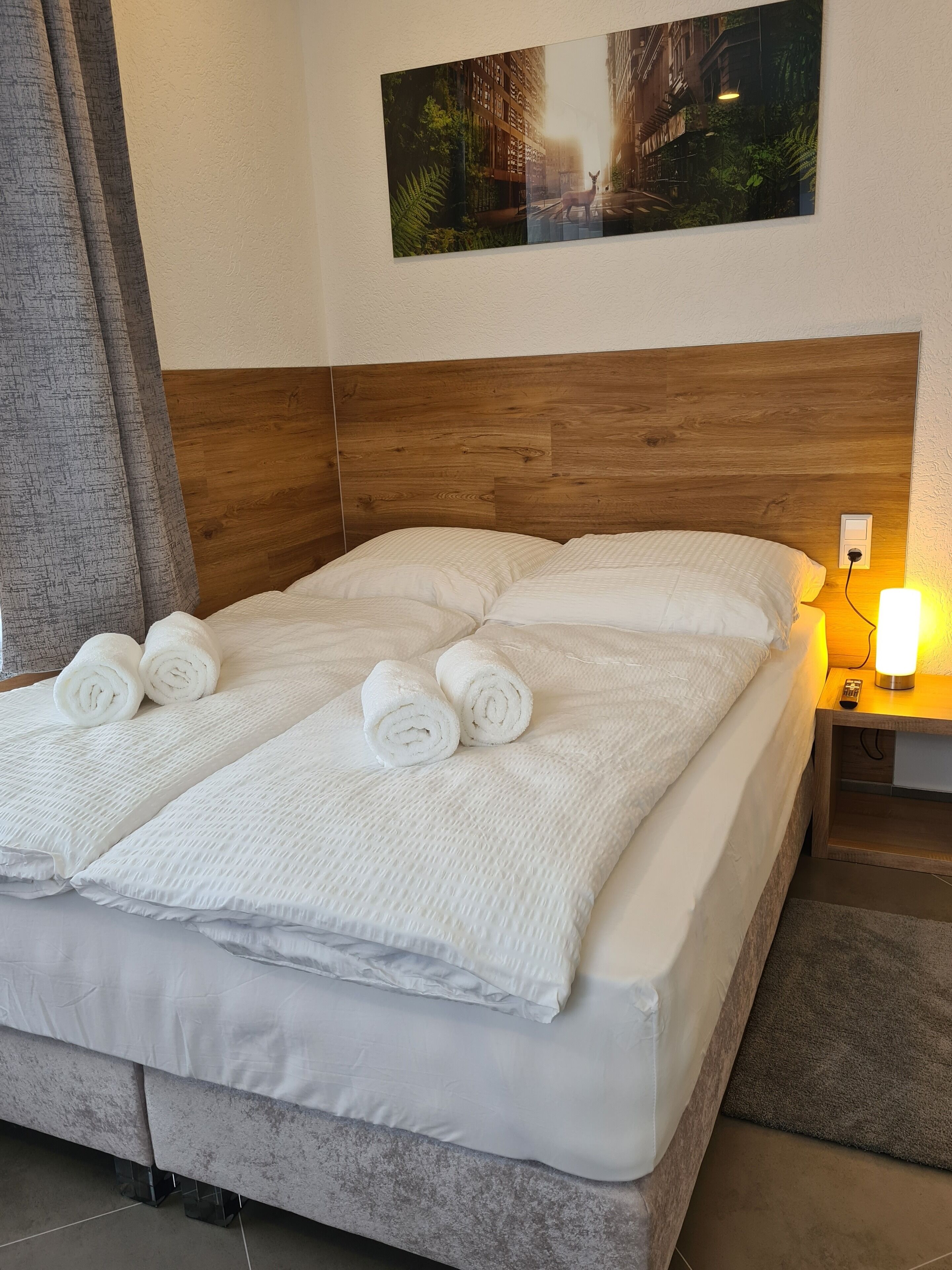Un lit bien fait, avec du linge de lit blanc et des serviettes pliées, une tête de lit en bois, une table de chevet avec une lampe et un tableau accroché au mur.