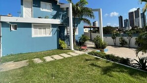 A residential building with a blue exterior, a small front yard with grass and a pathway, and a cityscape in the background. - Pousada Encanto dos Mares (Natal)