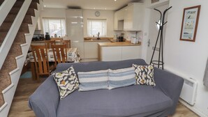 Ein Wohnzimmer mit grauem Sofa, gemusterten Kissen, einem Essbereich mit Tisch und Stühlen, einer Küchenzeile und einer Treppe.