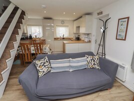 Una sala de estar con un sofá gris, cojines estampados, una zona de comedor con mesa y sillas, una cocina y una escalera.