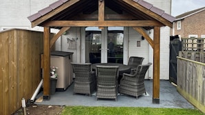 Ein überdachter Patio-Bereich mit Korbmöbeln, einer Holz-Pergola und einem weißen Backsteinhaus im Hintergrund.