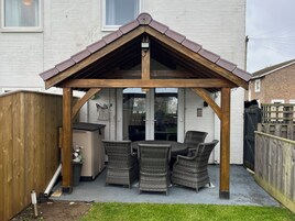 Un espacio cubierto con mobiliario de mimbre, un pérgola de madera y un edificio de ladrillo blanco al fondo.