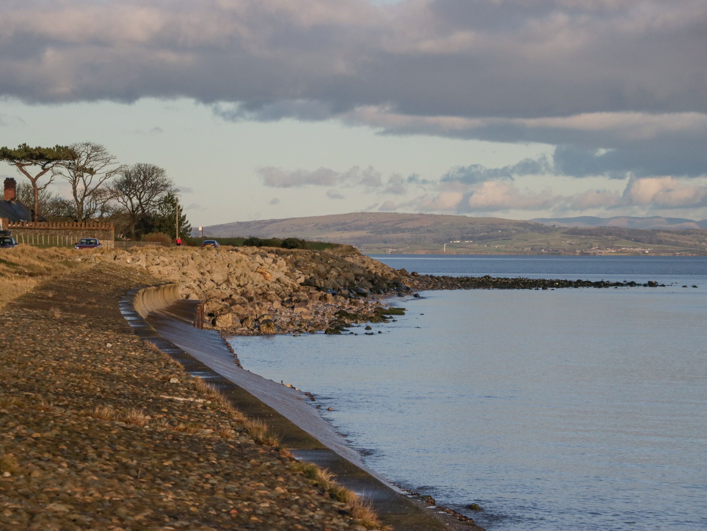 A coastal road with a rocky embankment, a paved walkway, and a view of the sea and distant hills.
