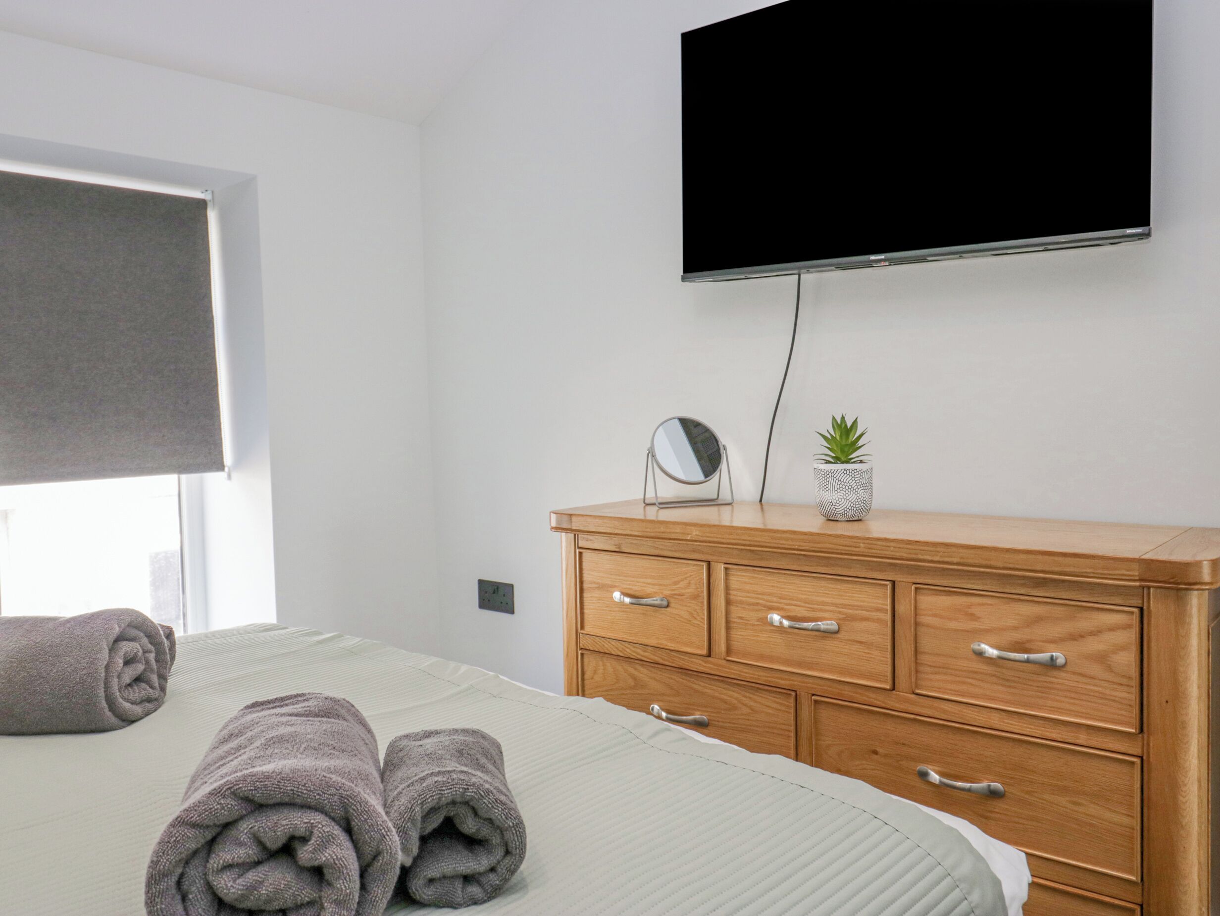 Un lit bien fait avec des serviettes pliées, une commode en bois avec des tiroirs, une petite plante en pot, un miroir rond et un téléviseur fixé au mur.