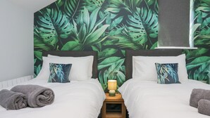 A bedroom with two beds, white bedding, grey towels, a nightstand with a lamp, and a tropical leaf wallpaper. - Crag View Cottage (Barrow-in-Furness)