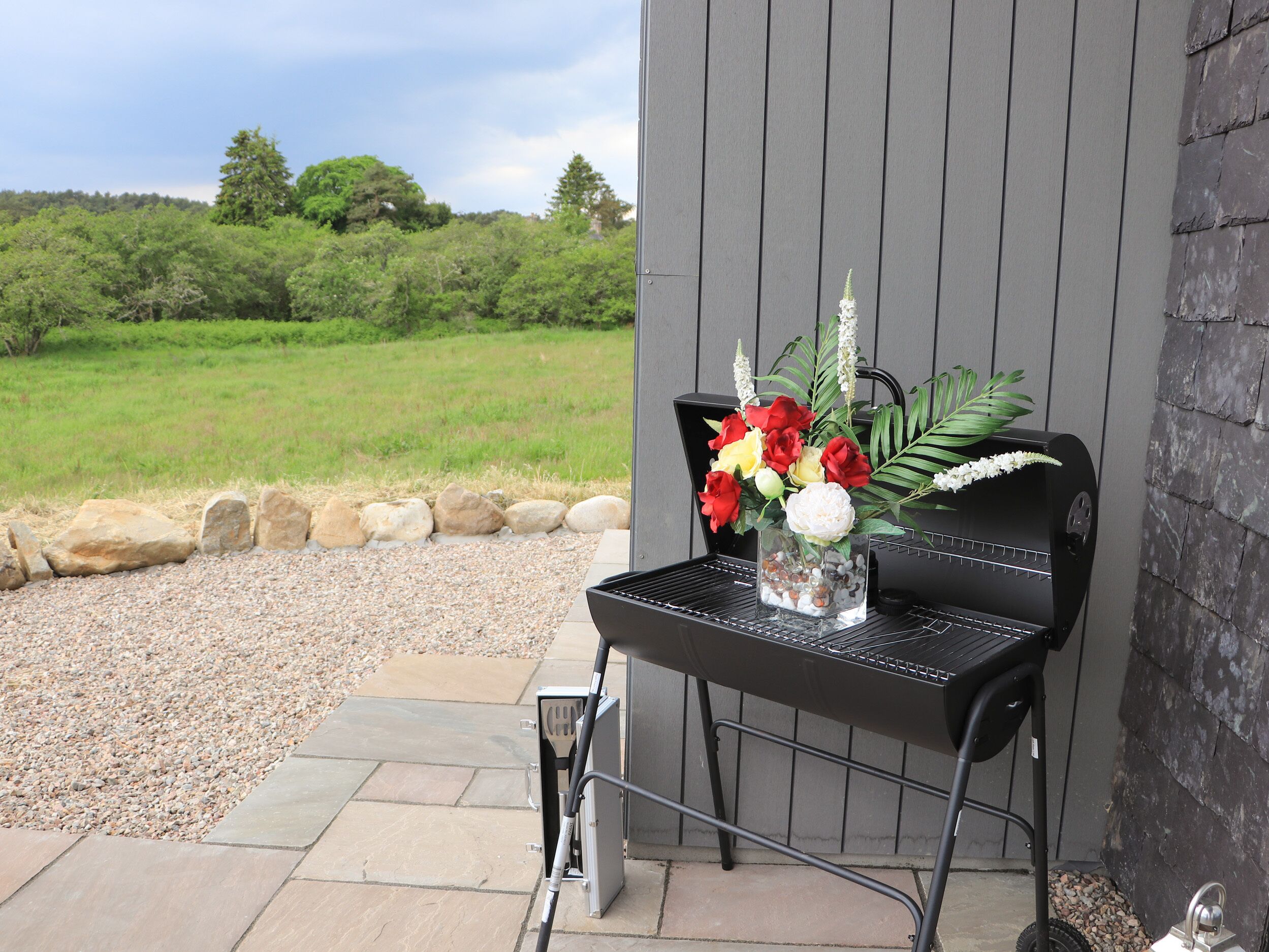 Eine Terrasse mit Grill, einem kleinen Tisch mit Blumenvase und Blick auf ein grünes Feld.