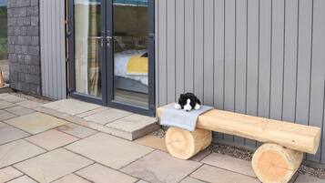 Un banco de madera con asiento de tronco y un peluche encima, ubicado frente a un edificio con puerta de vidrio y pared exterior gris.