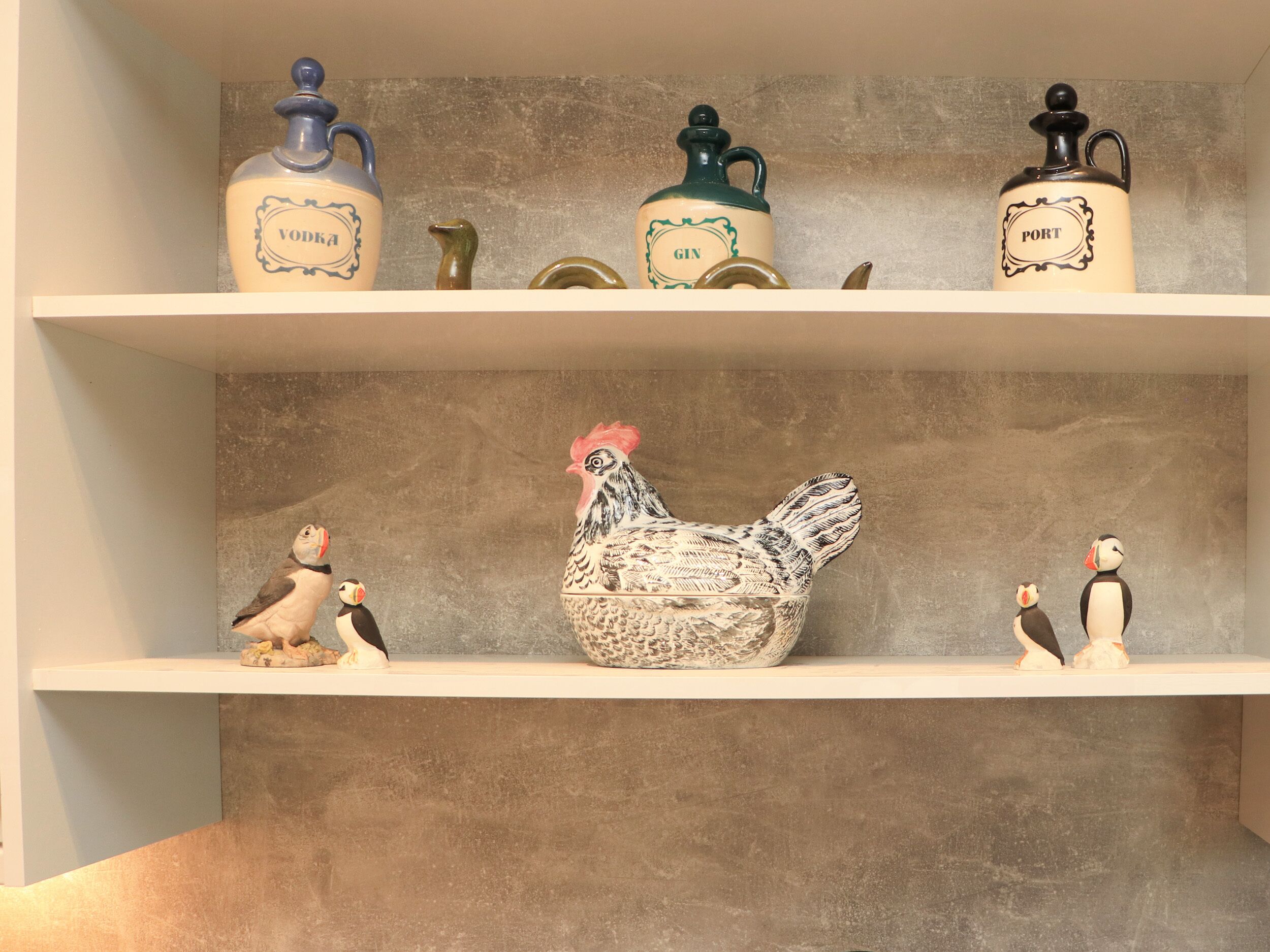 Een plank met decoratieve containers met de tekst 'VODKA', 'GIN' en 'PORT'. Daaronder staan keramische beeldjes van een haan en pinguïns.