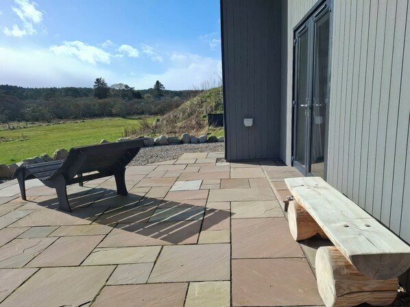 A patio with a bench, a wooden log, and a building with a door. - Red Squirrel (Dornoch)