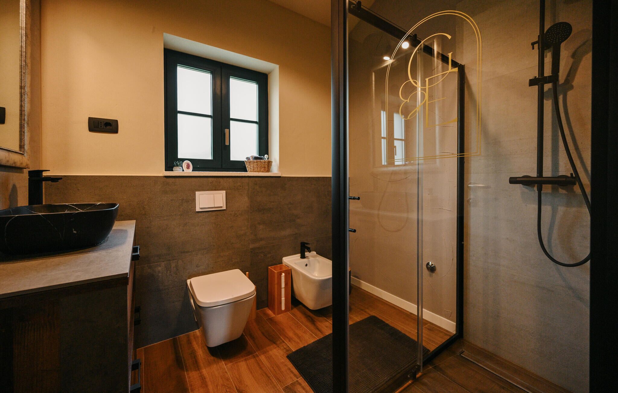 A modern bathroom with a glass shower enclosure, a black basin, a white toilet, and a window above the sink.