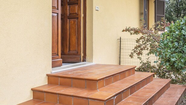 Una serie di gradini in terracotta che conducono a una porta in legno, un piccolo giardino con recinzione in filo di ferro e un muro giallo.