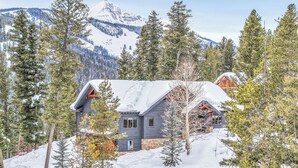 Une cabane aux fondations en pierre et aux murs en bois, entourée de neige et d’arbres, avec une montagne en arrière-plan.