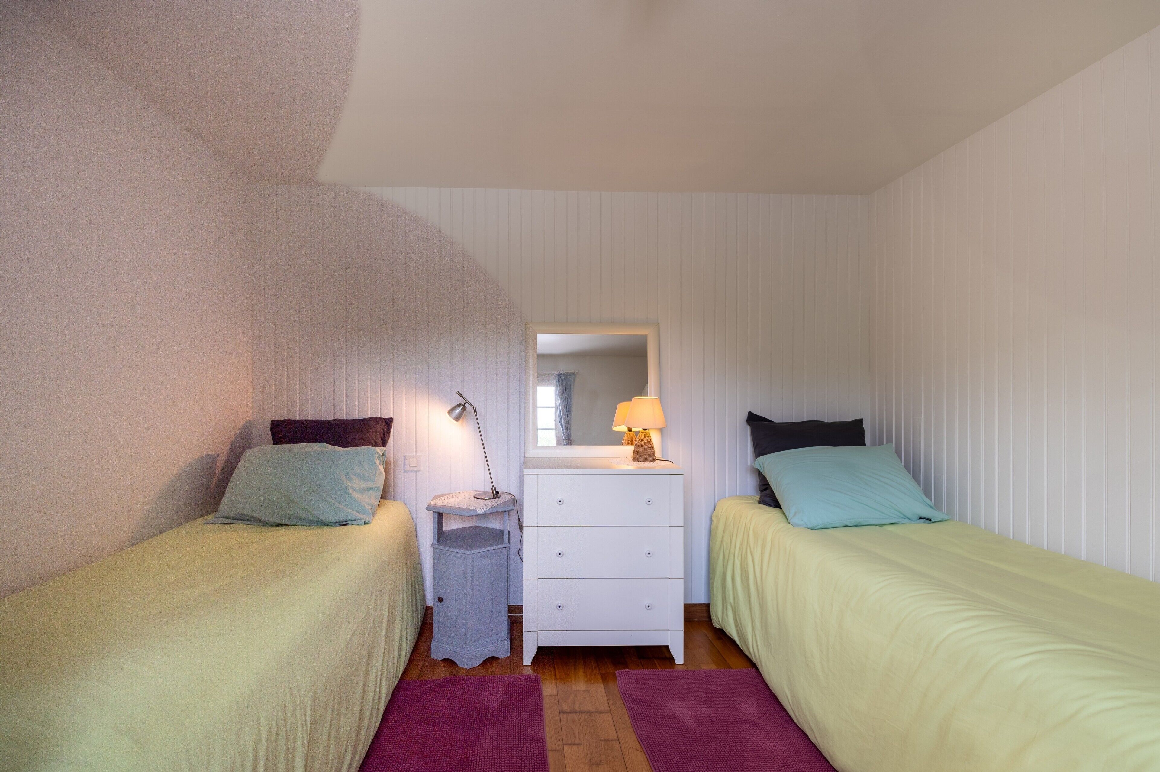 A small, single-room accommodation with two single beds, a bedside table with a lamp, a dresser, and a mirror.