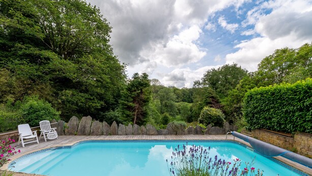A swimming pool surrounded by a wooden deck, with white lounge chairs and a stone wall.