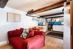 Un salon chaleureux avec un canapé rouge, des poutres en bois, une cuisine à l’arrière-plan et un tableau encadré au mur.